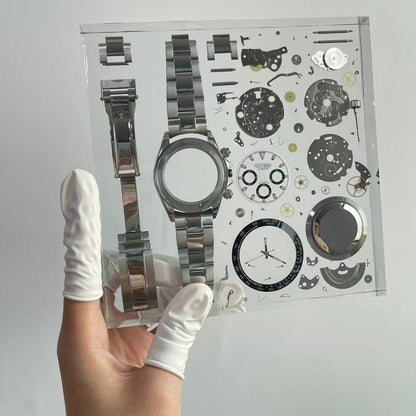 Hand holding a transparent resin block with watch components and a silver wristwatch.